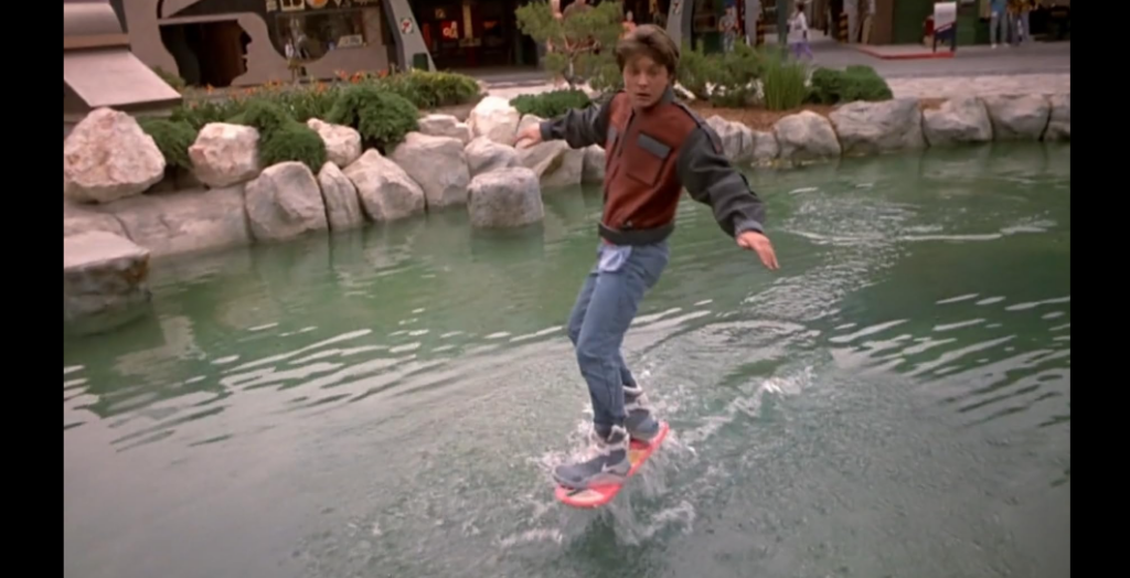 A young person rides a hoverboard over a pond, balancing with arms outstretched. They are dressed in a red and gray jacket, jeans, and sneakers. The background shows a rocky border around the water and a few people and a building in the distance.