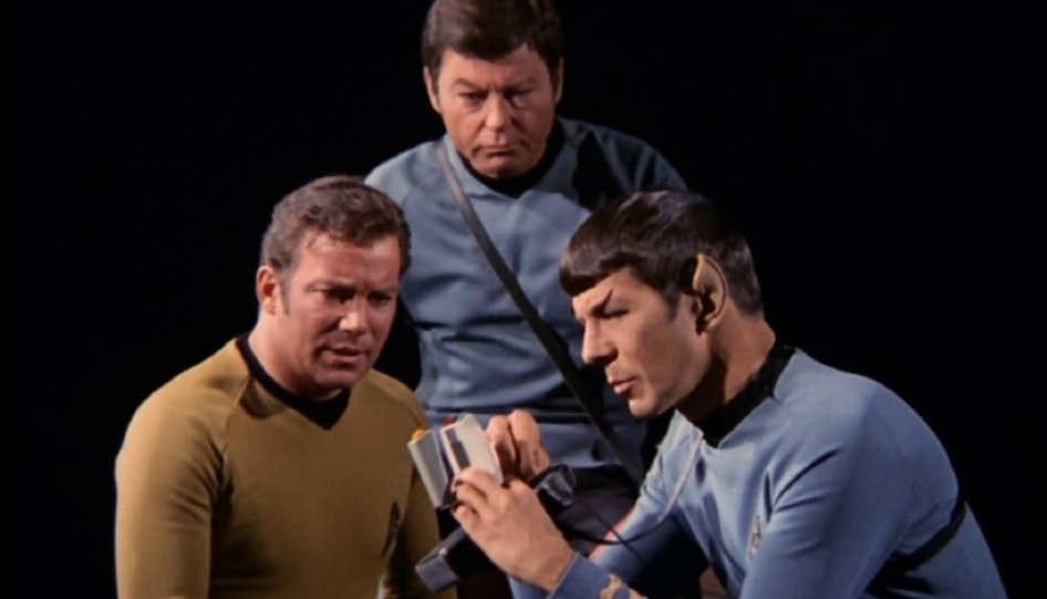 Three men in futuristic uniforms examine a handheld device. The man on the left wears a mustard-colored shirt, the man in the center wears a blue shirt, and the man on the right, with pointed ears, wears a blue shirt and focuses intently on the device.