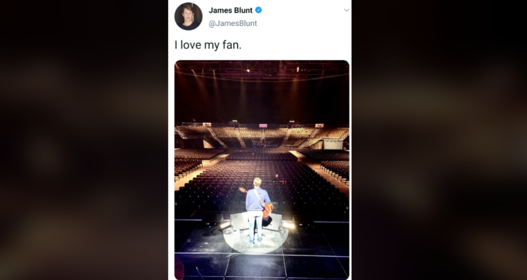 A person stands on a stage in a large, empty auditorium holding a guitar, facing the rows of empty seats. Above the image, there is a tweet from James Blunt that says, "I love my fan.