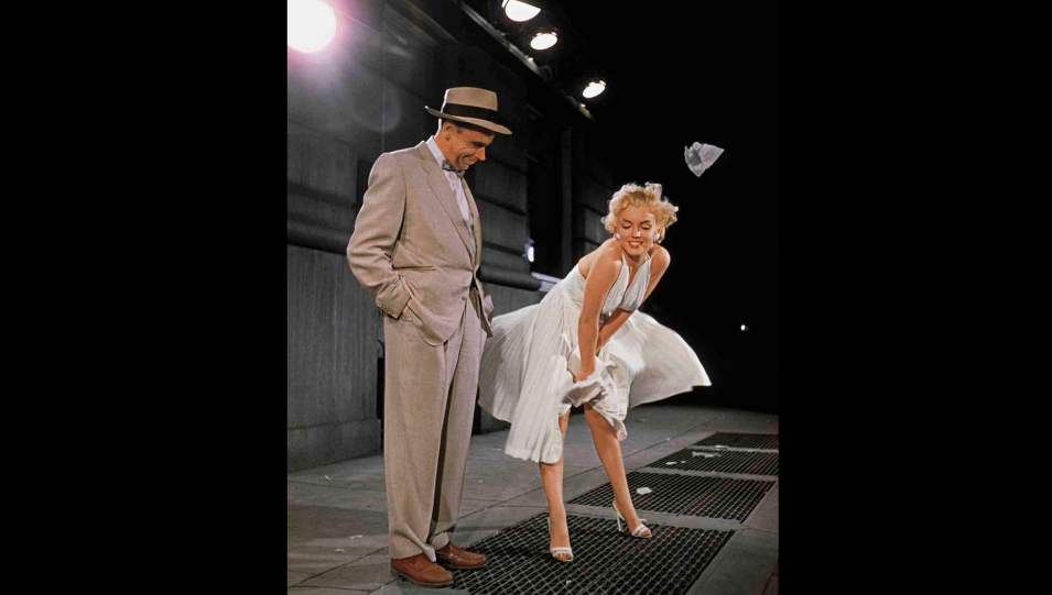 A woman in a white dress stands on a subway grate, smiling as the air blows her dress upwards. She holds down the dress to prevent it from flying up. A man in a beige suit, hat, and brown shoes stands beside her, looking at her with a smile. The background is dimly lit.