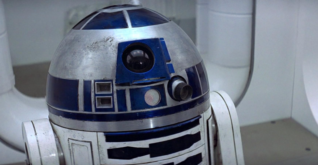 An image of a blue and white astromech droid with a cylindrical body, dome-shaped head, and various mechanical details, set against a backdrop of a sleek, white, futuristic interior.