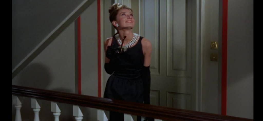 A woman in an elegant black dress with an elaborate pearl necklace and long black gloves stands on a balcony, looking upwards with a smile. She appears to be in a well-decorated indoor space with red accents on the walls and a staircase railing in the foreground.