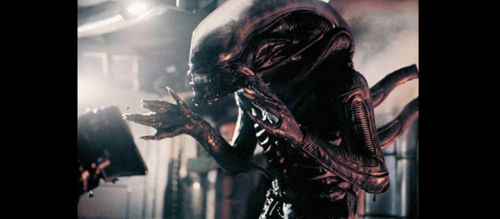 An image shows a detailed, dark-colored extraterrestrial creature from the movie "Alien." It has an elongated head, ridged body, and sharp claws, standing in a dimly lit, industrial-looking environment. The creature appears menacing, with a sinister posture.