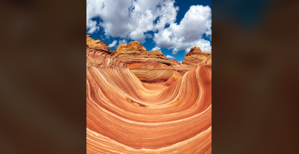 A stunning desert rock formation known as The Wave in Arizona, USA. The sandstone rocks display elegant, wavy patterns in shades of orange and red, contrasting against a bright blue sky with scattered white clouds.