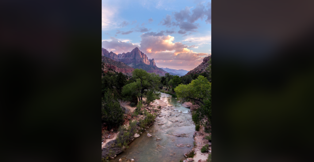 A serene river flows through a lush green valley surrounded by rocky mountains under a partly cloudy sky with a colorful sunset. Some peaks in the background are illuminated by the setting sun, creating a beautiful contrast of light and shadow.