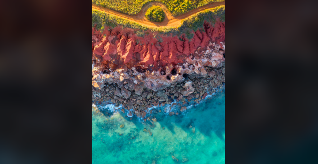 Aerial view of a coastal landscape showing a vivid contrast between red rocky terrain, a path cutting through green vegetation, and clear blue ocean water with small waves crashing onto the rocks.