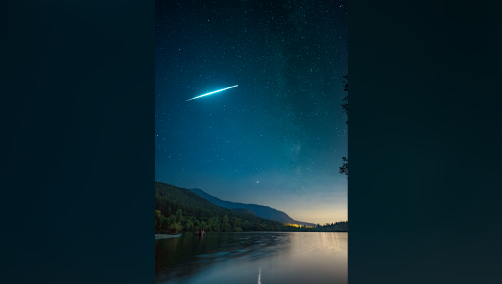 A bright shooting star streaks across a star-filled night sky above a serene lake. Silhouettes of mountains and trees frame the horizon, and the water mirrors the sky's myriad stars and the glowing meteor. Hints of distant lights are visible near the lake's edge.