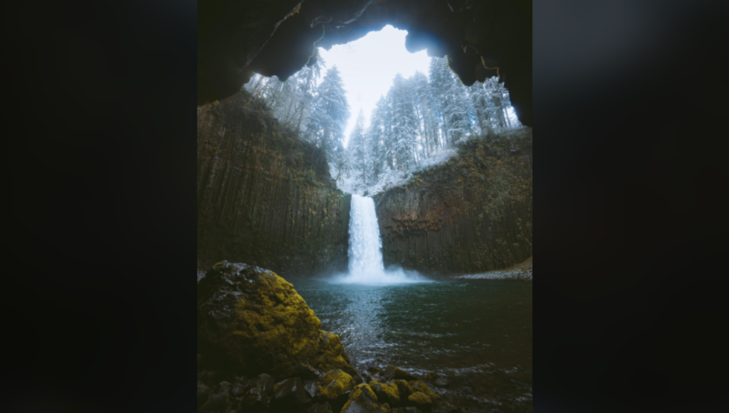 A stunning waterfall cascades into a serene pool surrounded by jagged cliffs. The photo is taken from the inside of a cave, framing the scene with moss-covered rocks in the foreground. Trees covered in light snow stand atop the cliffs under a pale blue sky.