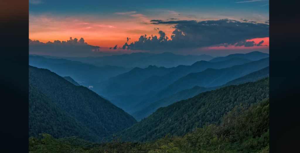 A serene mountain landscape at dusk, featuring layers of blue and green mountain ranges under a vibrant sky with orange, pink, and blue hues. Dense forests cover the mountains, and clouds float above the horizon.