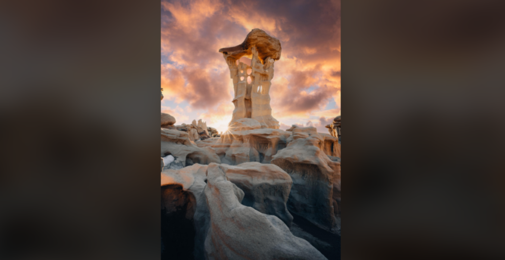 A surreal rock formation stands tall under a dramatic sky with pink and orange clouds at sunset. The unique and intricate shapes of the rocks create an otherworldly landscape, with smooth, eroded surfaces and distinctive, towering pillars.