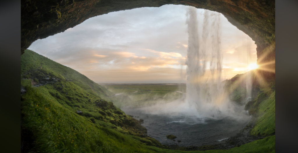 A view from behind a waterfall cascading into a pool below, with lush green hills surrounding the area. The sun is setting, casting a golden glow and creating a misty atmosphere. The sky is a mix of soft clouds and clear patches, adding to the serene landscape.