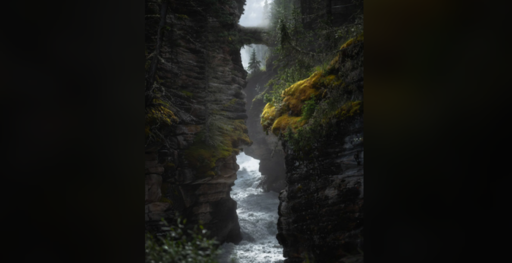 A misty, narrow canyon with steep, moss-covered rock walls. A rushing river flows through the bottom, and an ancient stone bridge spans the gorge overhead. Lush green vegetation clings to the rocks, adding a touch of color to the dark, mysterious scene.