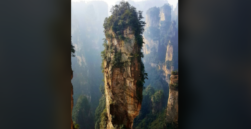 A tall, narrow sandstone pillar covered with green vegetation rises dramatically from the valley floor. Misty mountains and additional similar pillars are visible in the background, creating an otherworldly, serene landscape.