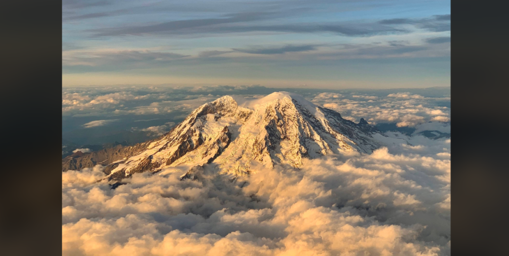 Aerial view of a snow-capped, twin-peaked mountain bathed in golden sunlight. The peaks rise above a sea of fluffy clouds under a partly cloudy sky. The mountain's rugged terrain contrasts with the softness of the surrounding clouds.