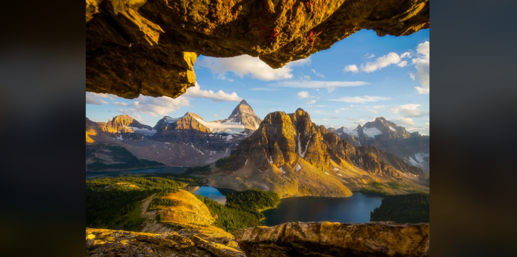 A stunning landscape view of a mountainous region during sunset, with rugged peaks illuminated by golden sunlight. A calm lake and dense evergreen forest are visible in the valley below, framed by rocky overhangs in the foreground, with a clear, partly cloudy sky above.