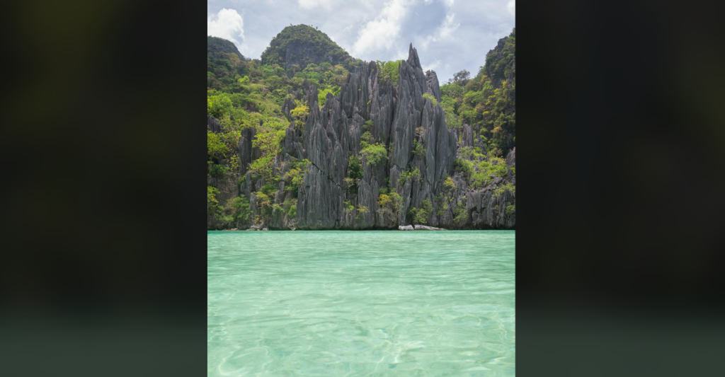 A stunning landscape featuring towering limestone cliffs with lush green foliage, set against a background of a partly cloudy sky. The foreground showcases clear, turquoise water, creating a picturesque tropical scene.