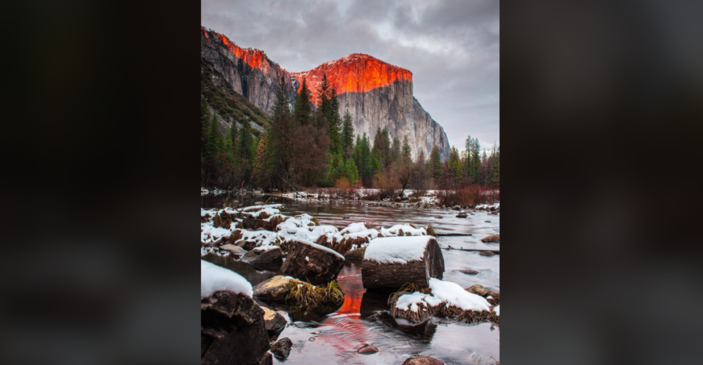 A serene river flows through a rocky landscape with snow on the rocks and trees along the bank. In the background, a majestic mountain peak glows with a vibrant red hue under a cloudy sky.