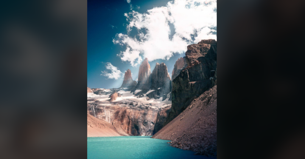 A stunning landscape featuring a turquoise lake in the foreground, surrounded by rugged, rocky terrain. In the background, there are towering sharp mountain peaks under a partly cloudy blue sky. The scene is illuminated by bright daylight.