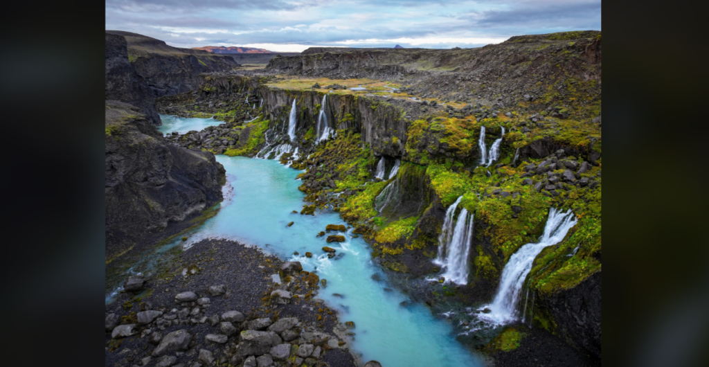 An aerial view of a valley featuring multiple waterfalls cascading down moss-covered cliffs into a winding turquoise river. The surrounding landscape is rugged with a mix of green vegetation and rocky terrain under a cloudy sky.