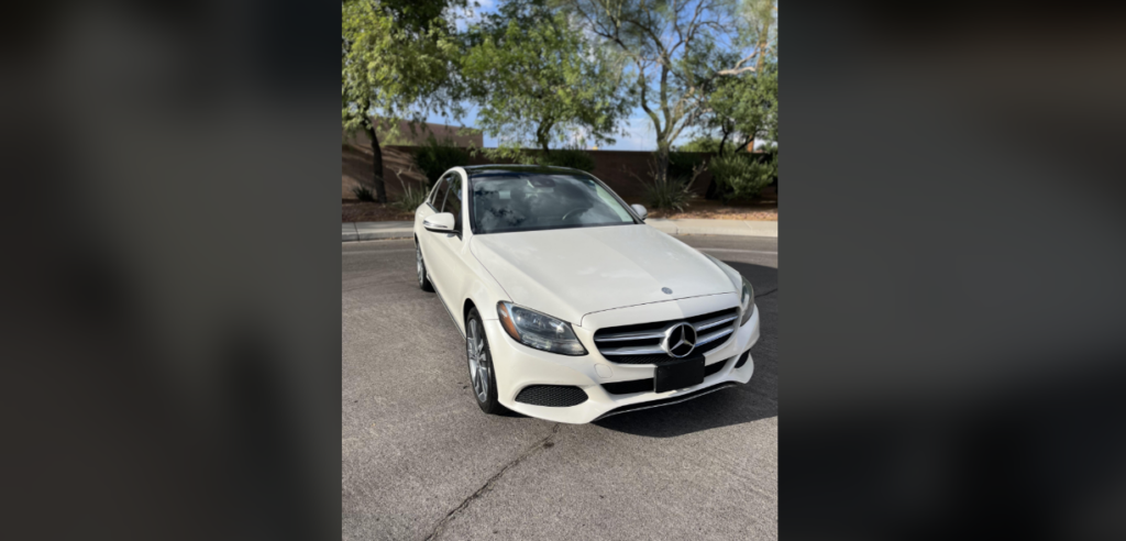 A white Mercedes-Benz car is parked in an outdoor setting on a paved surface. The background features trees, shrubs, and a brown wall. The car is facing forward, showcasing its grille and front features.