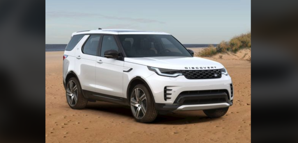 A white Land Rover Discovery is parked on a sandy beach with grassy dunes in the background. The SUV has a sleek design and appears modern and sturdy, ideal for off-road adventures. The sea is visible in the distance under a partly cloudy sky.