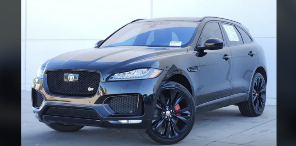 A sleek black Jaguar F-Pace SUV parked outdoors on a concrete surface. The vehicle features a glossy finish, tinted windows, and black alloy wheels with red brake calipers. Visible behind the car is a white wall, indicating an outdoor or showroom setting.