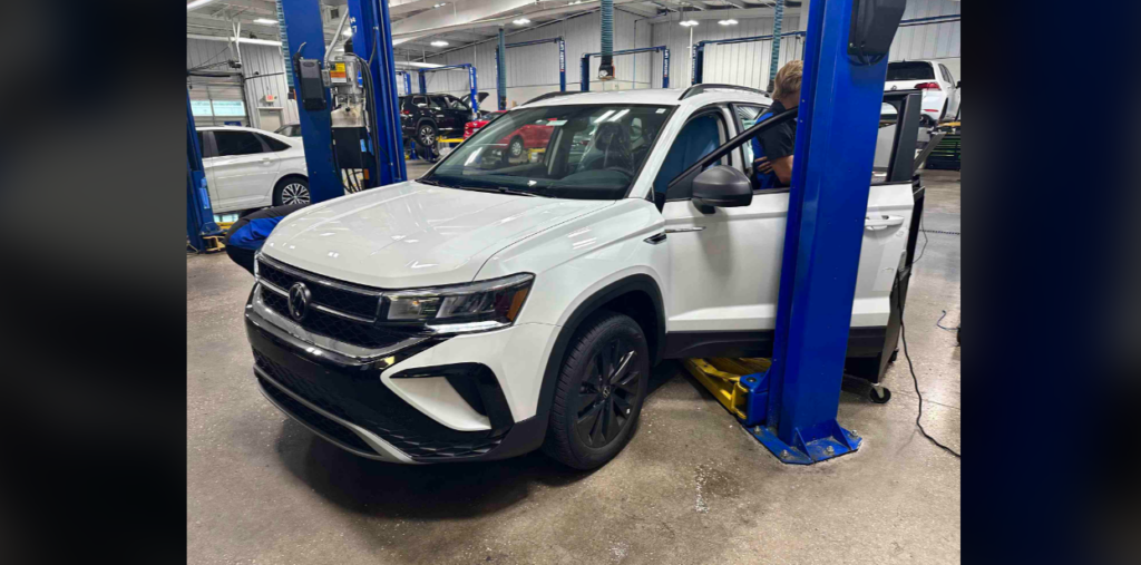 A white SUV is being serviced in an automotive repair shop. The vehicle is lifted slightly off the ground by a hydraulic lift, and a person is inspecting or working on it through the open driver's side door. The garage is spacious with other cars in the background.