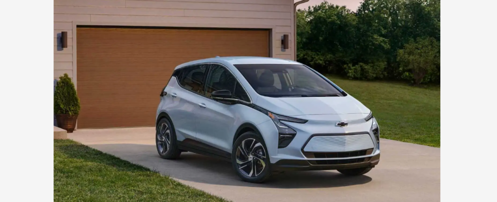 A light blue Chevrolet electric vehicle is parked on a driveway in front of a brown garage door. The car features a modern, sleek design with a closed front grille, black trims, and distinctive alloy wheels. Lush greenery surrounds the house in the background.