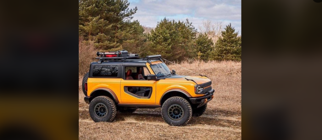 A bright orange off-road SUV with black accents is parked on a dirt path surrounded by grass and trees. The vehicle has large tires, roof-mounted equipment, and open panel doors, emphasizing its rugged design. The sky is partly cloudy in the background.