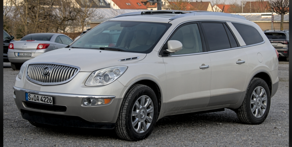 A silver Buick Enclave SUV is parked in a gravel lot. The vehicle has a chrome front grille, roof rails, and tinted windows. Other cars and buildings are visible in the background.