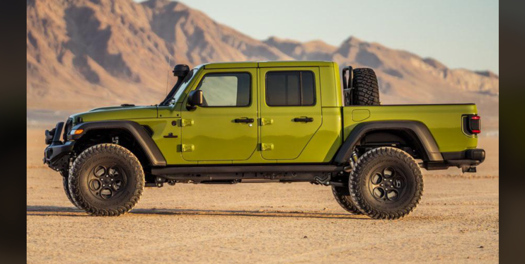 A green off-road truck with large tires is parked in a desert setting with mountains in the background. The vehicle has a rugged appearance and appears ready for an adventure across the sandy terrain.