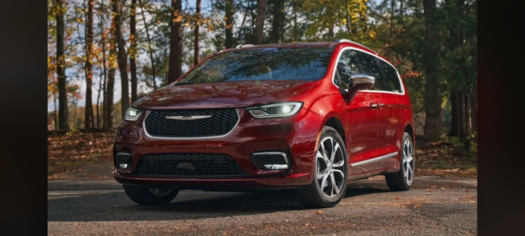 A red minivan is parked on a road in a wooded area. The vehicle has sleek, modern styling with a prominent front grille, chrome accents, and large wheels. Trees with autumn foliage are visible in the background.