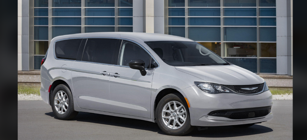 A silver minivan is parked on a paved surface in front of a building with large blue-tinted windows. The van has tinted rear windows, a sleek design, and five-spoke wheels. The building in the background has a modern glass and concrete exterior.