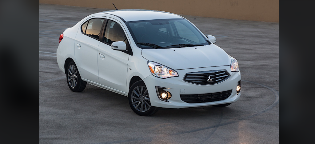 A white compact sedan car is parked on a gray concrete surface. The car features a sleek design with chrome accents on the front grille, fog lights, and alloy wheels. The background is plain and unobtrusive, focusing attention on the vehicle.