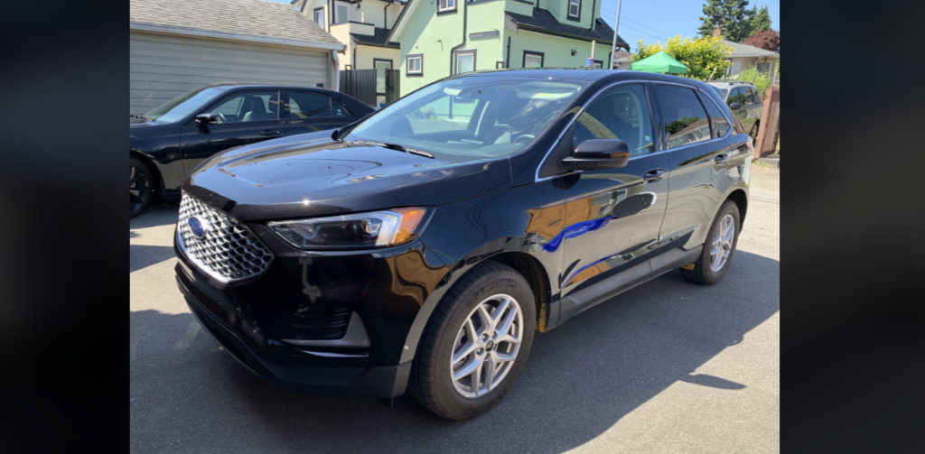 A black SUV is parked on a driveway in front of a residential house. The vehicle has a shiny exterior and silver alloy wheels. There are other cars and houses visible in the background under a sunny sky.