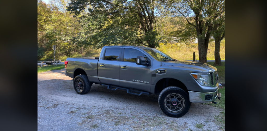 A silver Nissan Titan XD pickup truck is parked on a gravel road. The truck is in a rural area with trees and green foliage in the background, and sunlight is shining through the trees. The truck has dark-tinted windows and black side steps.