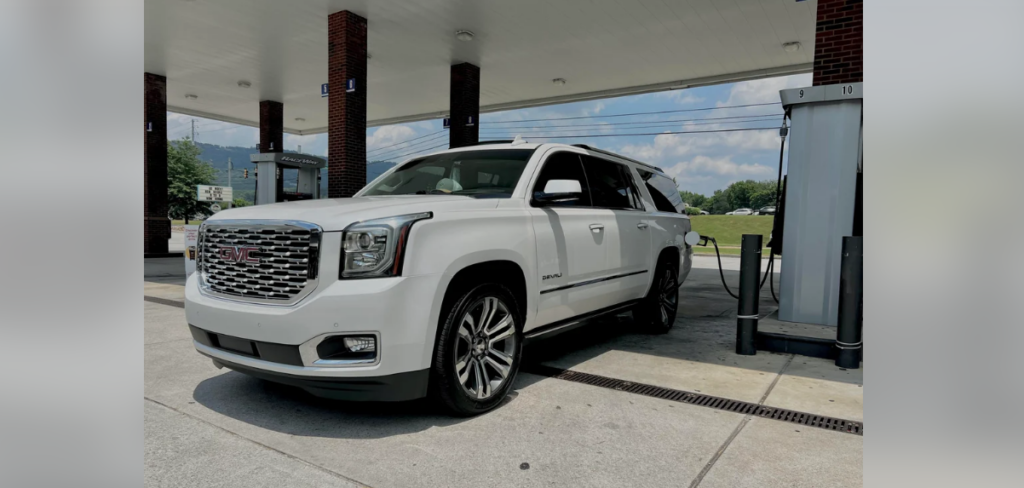 A white GMC Yukon is parked at a gas station under a canopy with red brick pillars. The driver's side door is closed, and the vehicle is positioned near a fuel pump. It's a clear, sunny day with a partly cloudy sky.