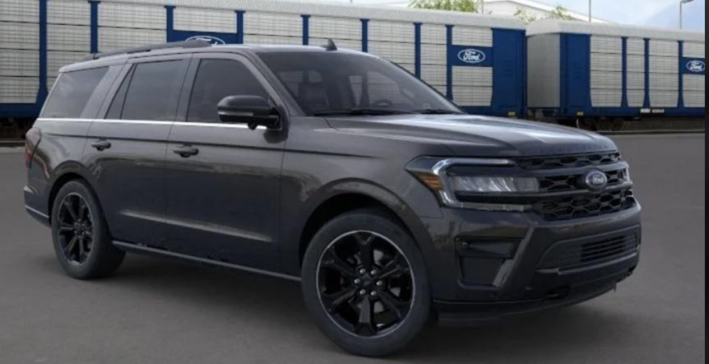 A black Ford SUV is parked on a paved surface with a Ford train in the background. The vehicle has a sleek design, black alloy wheels, and tinted windows. The front grille features the Ford emblem, and the overall look suggests a robust yet stylish appearance.