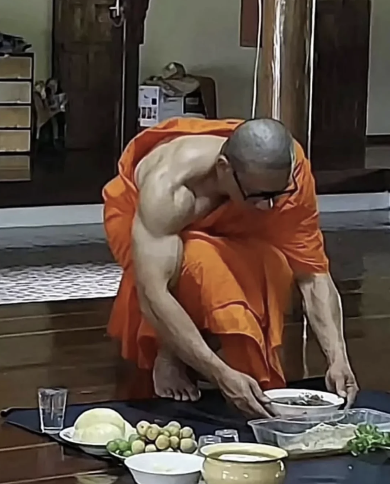 A bald man in an orange robe is kneeling on the floor and setting down a bowl. The scene is indoors with various food items placed on the floor in front of him, including vegetables and other dishes. A glass of water is also visible.