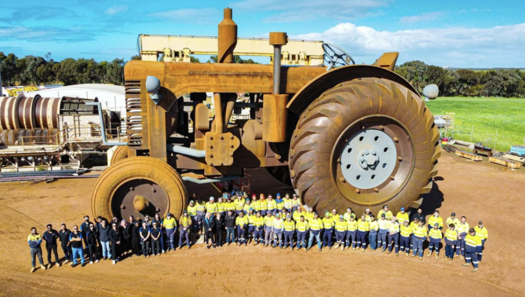 A large group of people dressed in uniforms stand together in front of a massive, rusted industrial machine with enormous wheels. The machine is set against a backdrop of a clear blue sky and green fields. Equipment and buildings are visible in the background.