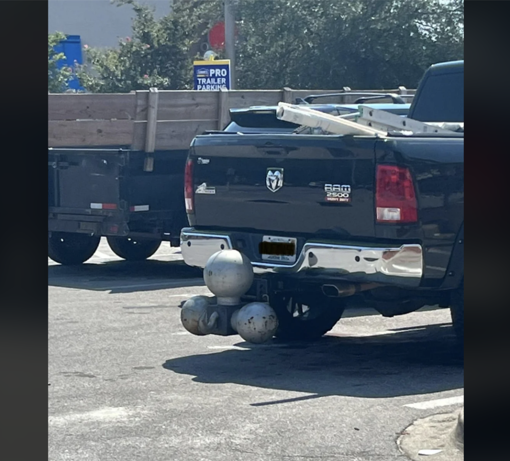 A black Ram 1500 truck is parked with a set of novelty large gray metallic spheres attached to its hitch. Another truck with a wooden cargo bed is visible to the left. A "Pro Trailer Parking" sign is partially seen in the background.