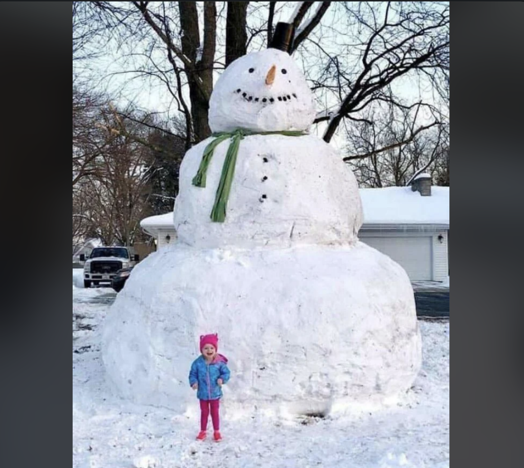 A young child in a pink hat and blue jacket stands in front of a giant snowman. The snowman, adorned with a green scarf, black buttons, and a carrot nose, towers over the child. Snow-covered trees and a house with a garage are visible in the background.