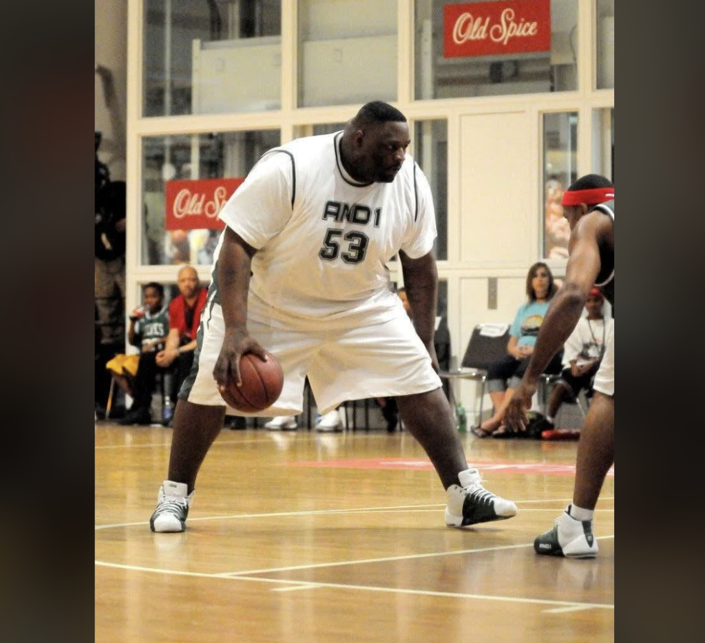 A basketball player wearing a white jersey with the number 53 dribbles the ball on an indoor court, facing a defender. Spectators are visible in the background, and advertisements for Old Spice can be seen on the walls.