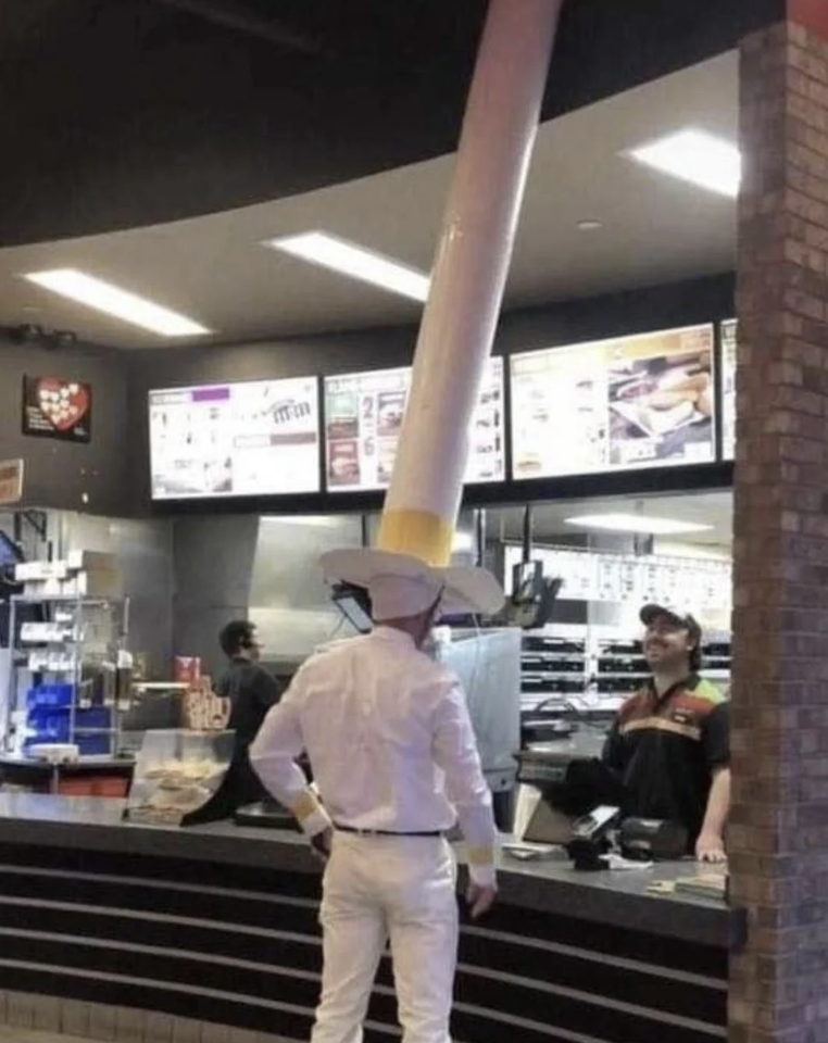 A person dressed in a cowboy outfit with a giant cigarette prop on their head stands at a fast-food counter. The employee behind the counter looks at them with an amused expression. The background includes menu boards and typical fast-food restaurant decor.