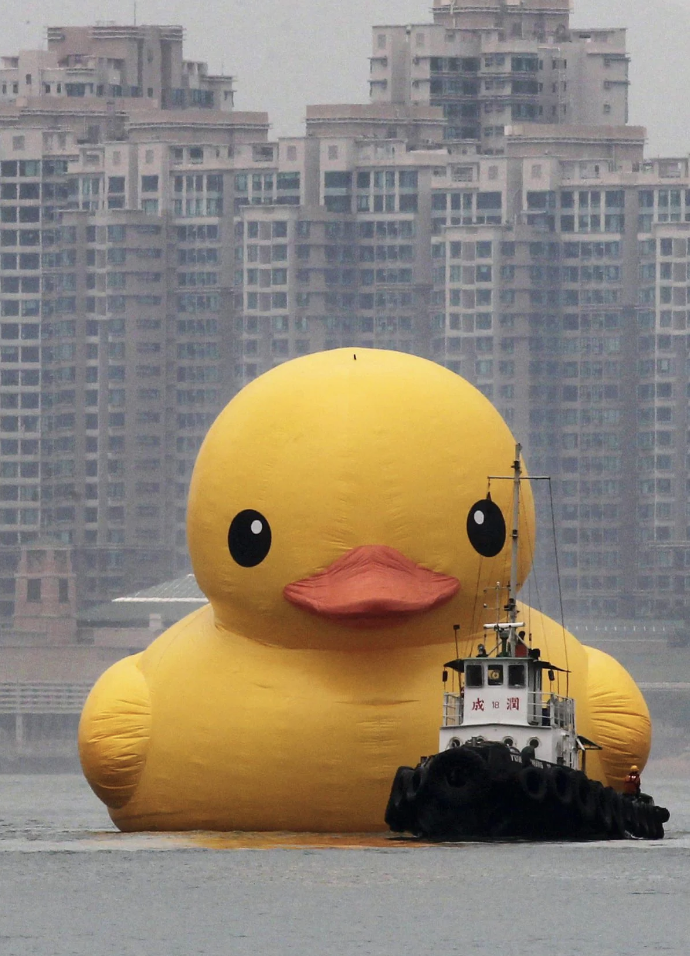 A large, yellow rubber duck float is positioned in the water near a cityscape with high-rise buildings. A small boat is closely accompanying the giant rubber duck, guiding it through the water. The sky is overcast.