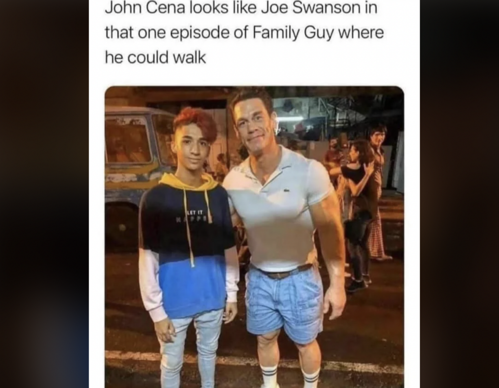 A young man with red hair wearing a blue and white hoodie and jeans stands next to a muscular man in a tight white t-shirt and blue shorts. They are posing for a picture on a street at night with people and a vehicle in the background.