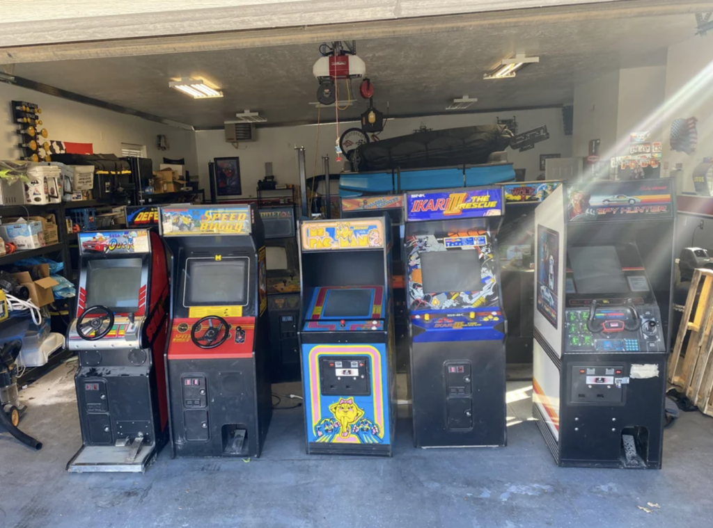 A garage with several vintage arcade game machines lined up side by side, including "Super Sprint," "Harley-Davidson & L.A. Riders," "Pac-Man," "Krazy Bowl," and "Operation Wolf." The space also contains various tools and equipment in the background.