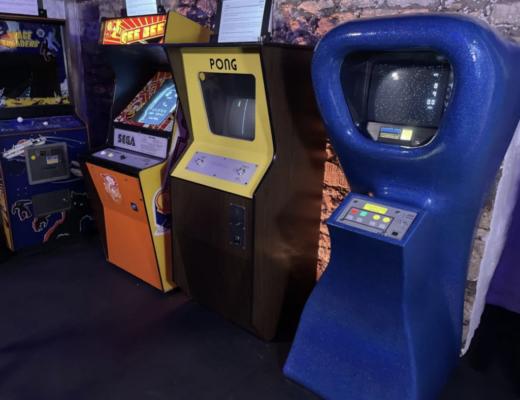 A row of vintage arcade game machines, including 'Space Invaders,' 'SEGA's Deep Scan,' 'Pong,' and 'Computer Space,' all housed in a dimly lit room with a rustic concrete wall in the background. The machines feature colorful designs and classic controls.