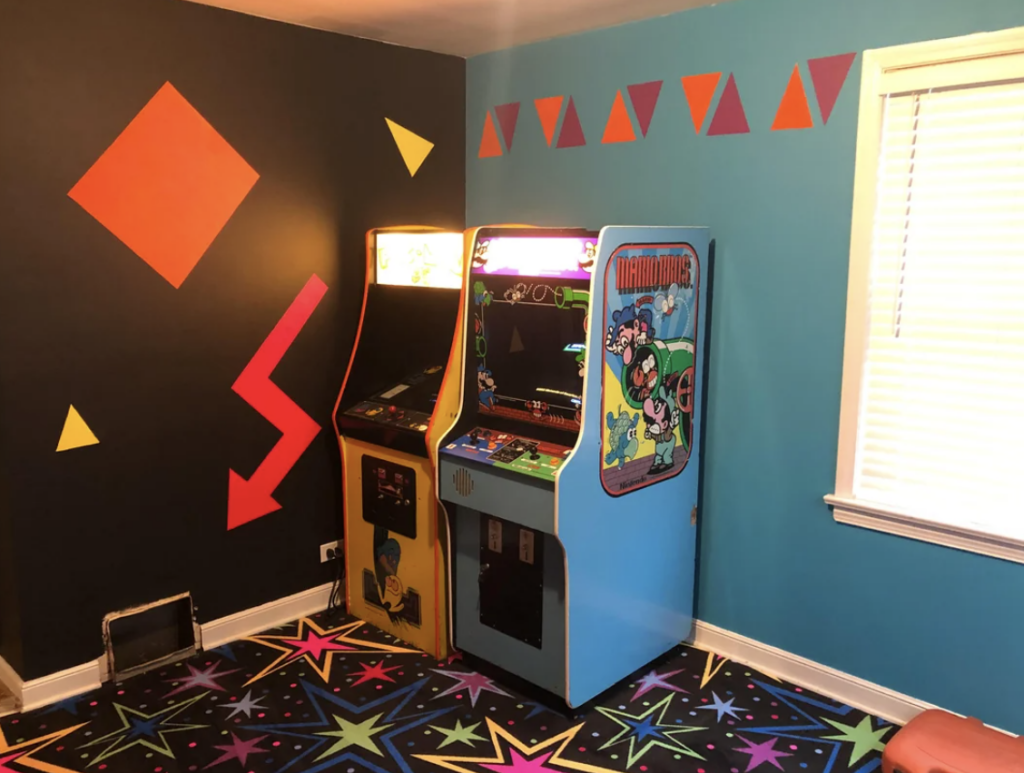 A room with colorful retro decor featuring two arcade machines against a wall. The wall is painted dark with geometric shapes. The carpet is vibrant with star patterns, and a window with white blinds lets in some light.