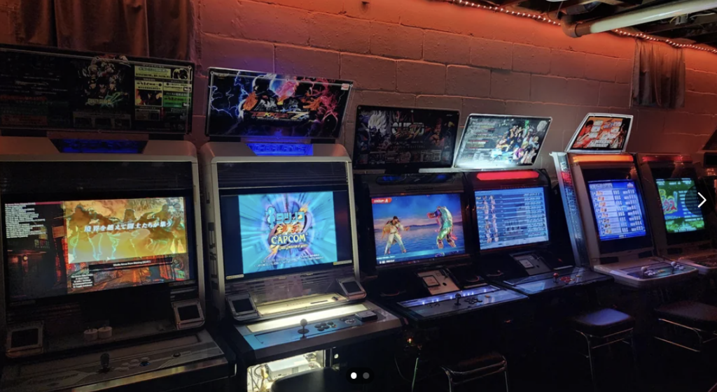 Dimly lit arcade with six vintage gaming machines side by side. Each screen displays a different game, with the central machine showing a "Capcom" logo. Stools are placed in front of each machine for players. The room is illuminated by red and white ceiling lights.